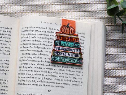 Magnetic bookmark featuring a stack of spooky books with a pumpkin on top. Names of book show on their spine. Colors are orange, green, and yellow.